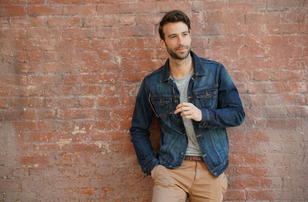 A smiling young man in a blue jean jacket leaning against a wall.