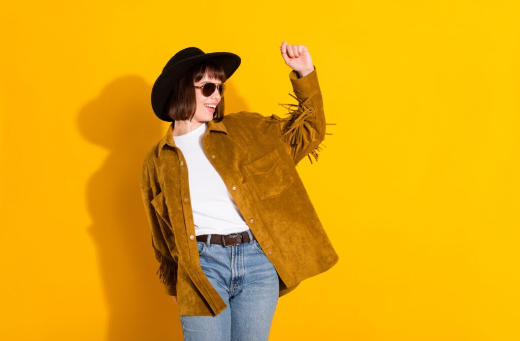 A woman wearing a black cowboy hat, a brown fringe jacket, a white shirt, and blue denim jeans. She's standing in front of a yellow background.