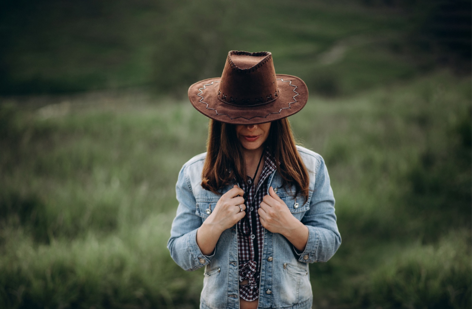 A woman wearing a brown cowboy hat with the brim tipped low to hide her face. She's pulling her denim jacket closed and wearing a checkered button up shirt.
