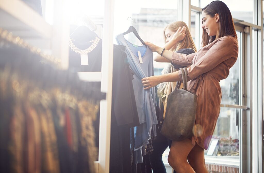 Two people shop in a sun-filled store, pulling out a blue dress to look at it more closely. Both people are smiling.