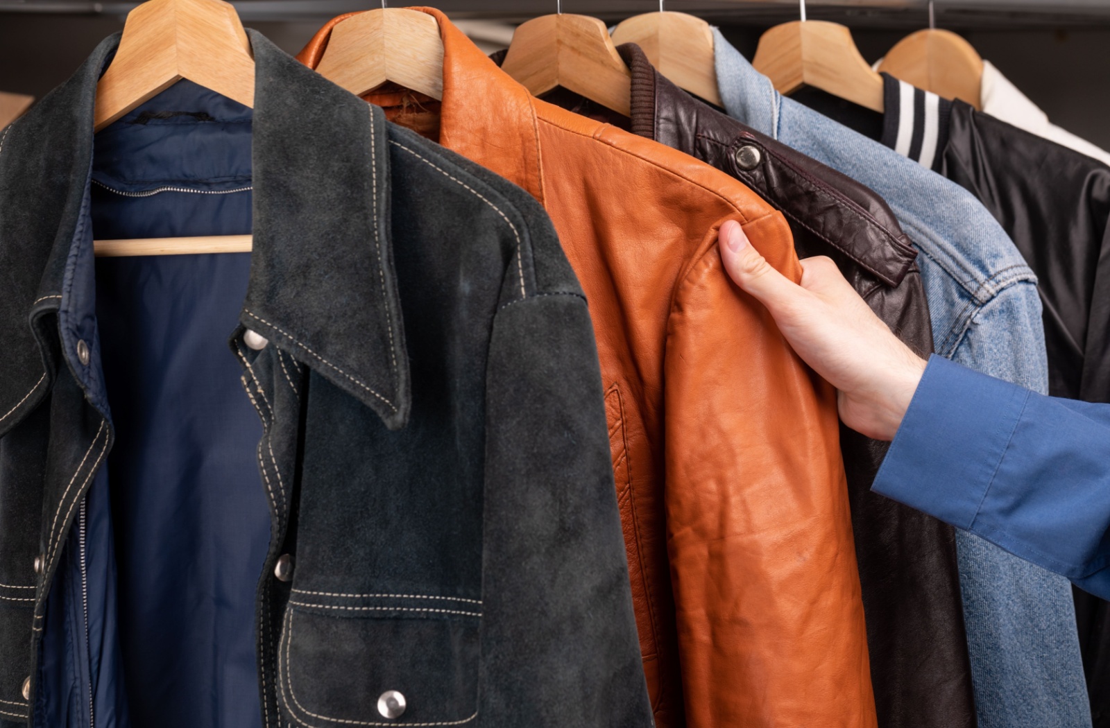 A hand touching a pre-loved brown leather jacket hanging on a rack of second-hand outerwear.