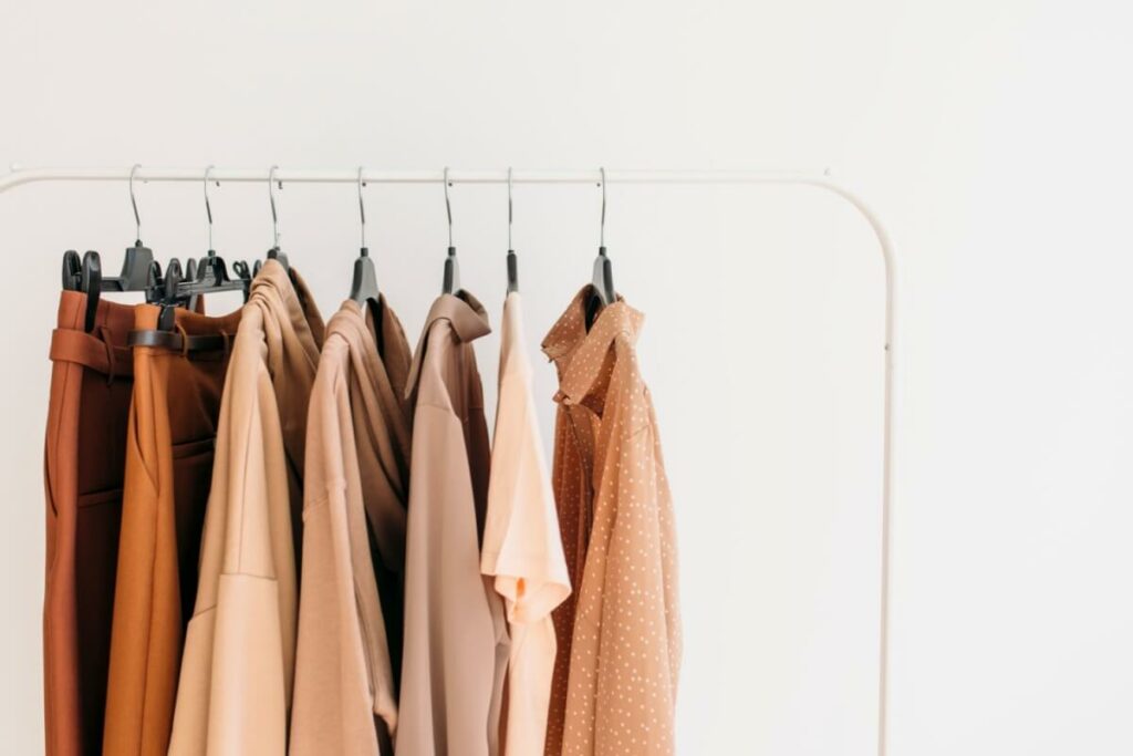 A selection of minimalist clothing in neutral earth tones hanging on a white garment rack against a white wall.