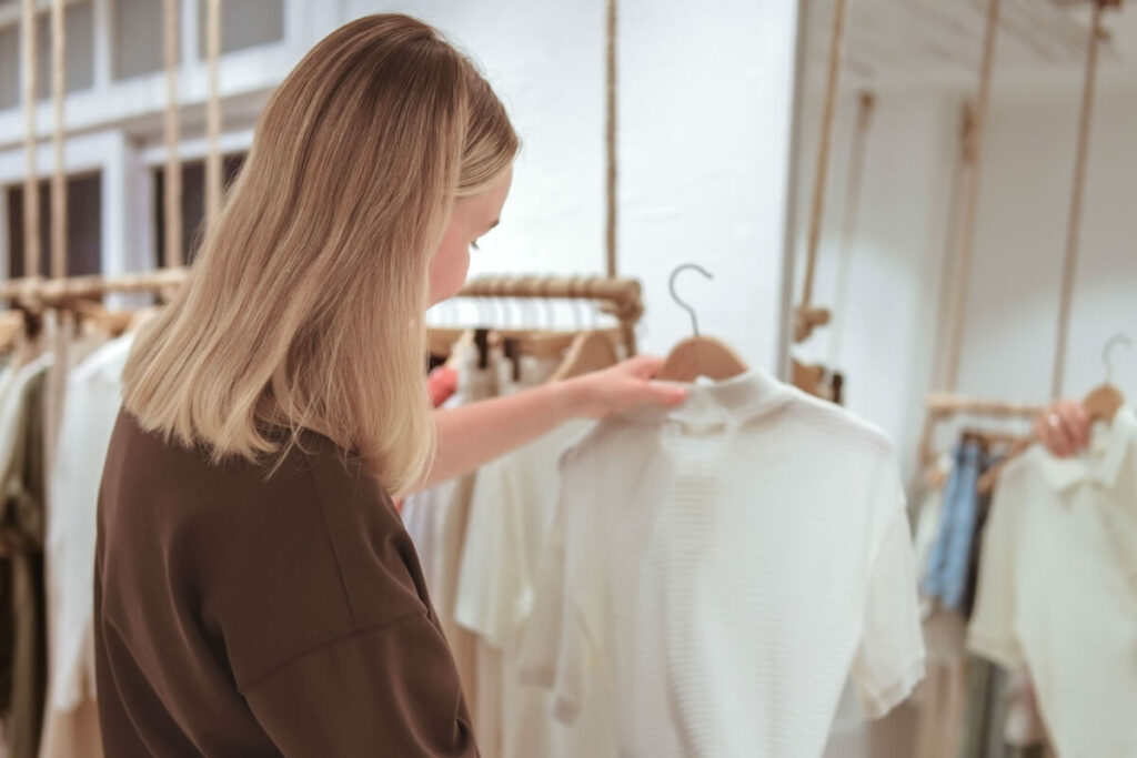 A person with blonde hair seen from behind browsing cream-colored tops on wooden hangers in a minimal fashion boutique.